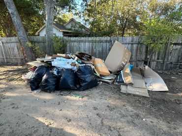 A pile of trash bags and cardboard boxes in a backyard area near a wooden fence.