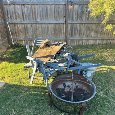 Outdoor fire pit with wooden chairs and a doormat on grass near a wooden fence.
