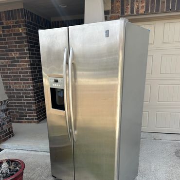 Stainless steel side-by-side refrigerator placed outdoors on a driveway.