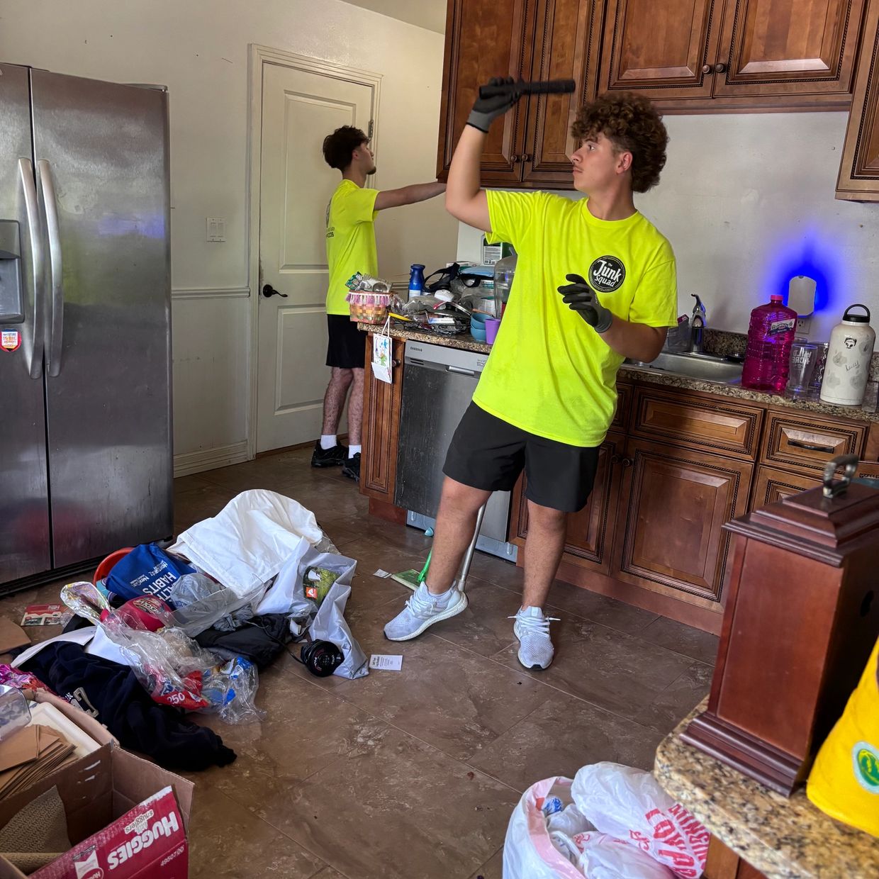 Two men in neon shirts cleaning a cluttered kitchen.