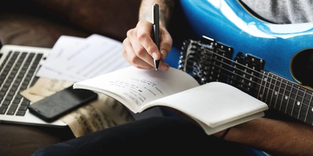 Person writing music notes in a notebook with a blue electric guitar and laptop nearby.