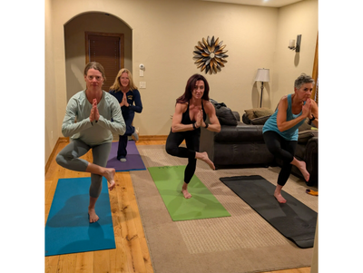 Four women practicing yoga balancing on one leg in a cozy living room.