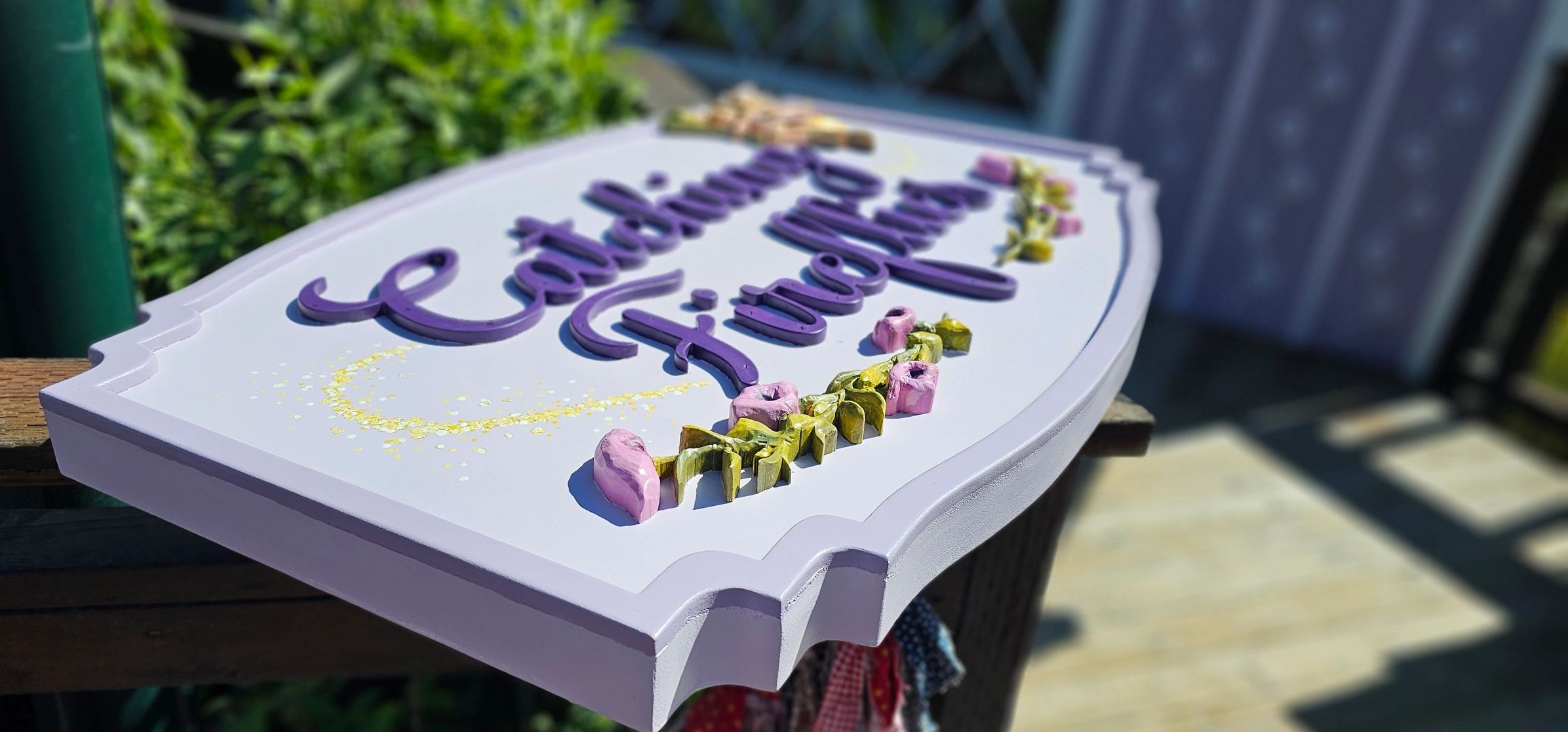 Decorative sign with purple text and floral accents on a wooden surface outdoors.