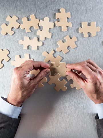 Hands assembling beige puzzle pieces on a gray surface.