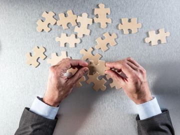 Hands assembling beige puzzle pieces on a gray surface.