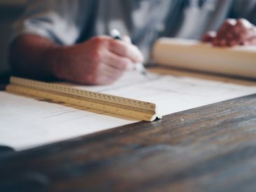 Person sketching architectural plans with a ruler on a wooden table.