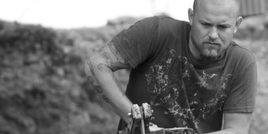 A man with a beard and tattooed arms works intently with a mechanical tool pumping water outdoors.