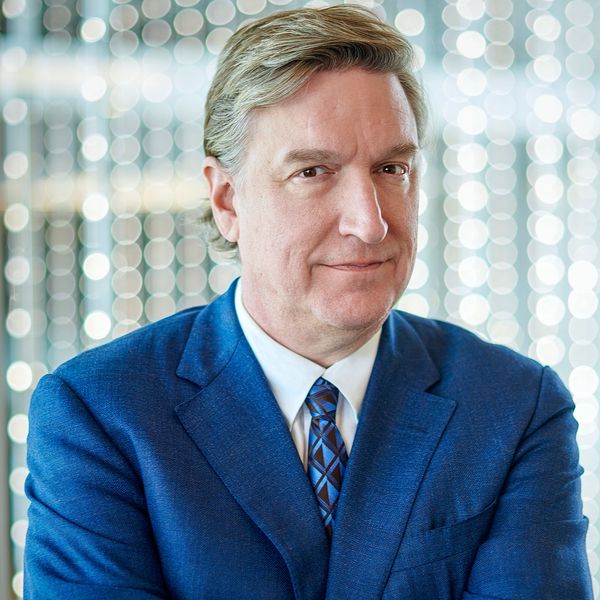 Man in a blue suit with arms crossed, standing in front of a light-filled background.