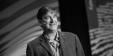 A man with short hair speaks onstage, smiling as he wears a blazer and microphone headset.