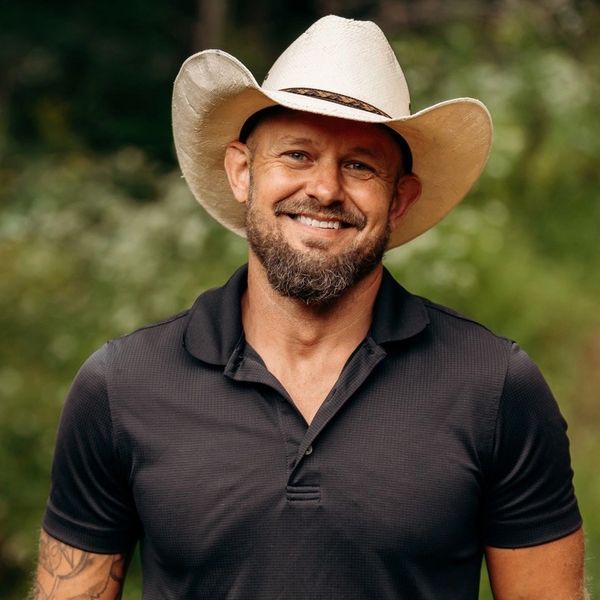 Man outdoors wearing a black polo and cowboy hat, smiling with hands in pockets.