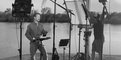 A man stands outdoors near the water, speaking on camera while a crew adjusts equipment around him.