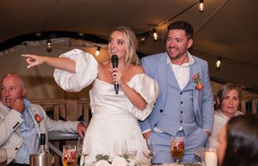 Bride in white dress pointing during wedding speech with groom beside her.