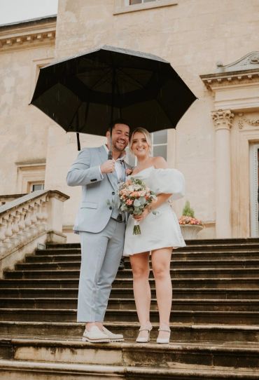 Smiling couple in wedding attire holding an umbrella on rainy steps.