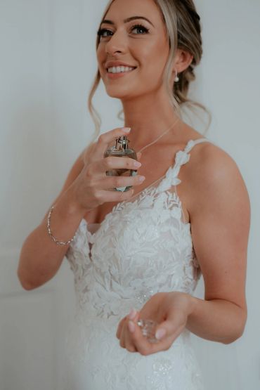 Woman in a white floral dress spraying perfume and smiling.
