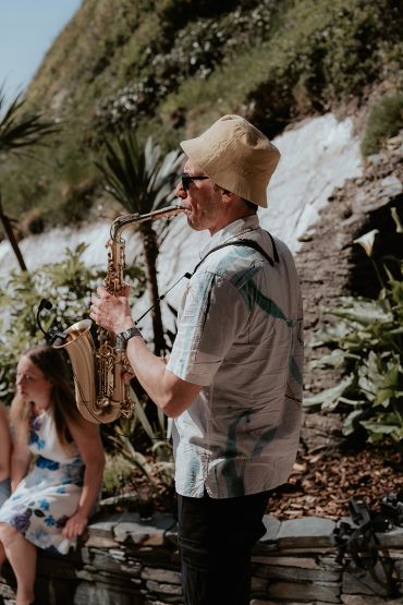 Man playing saxophone outdoors in casual summer attire.