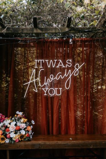 Neon sign reading "It was always you" with flowers below.