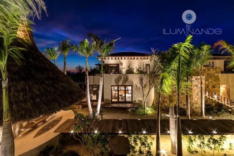 "Iluminación cálida destaca senderos, palapas y palmeras, creando un ambiente tropical elegante."