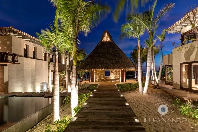 Iluminación cálida destaca senderos, palapas y palmeras, creando un ambiente tropical elegante
