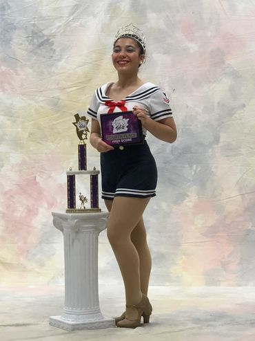 Young woman in a sailor costume holding a fourth place award, smiling beside a trophy on a pedestal.