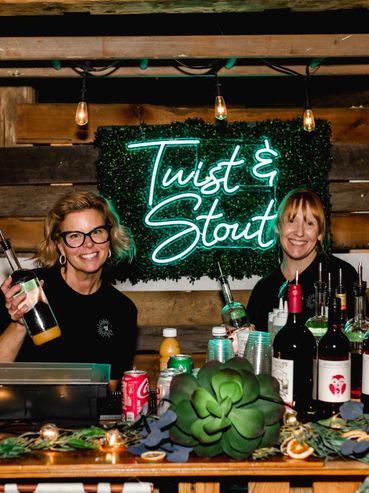 Two bartenders smiling behind a rustic bar with a neon "Twist & Stout" sign.