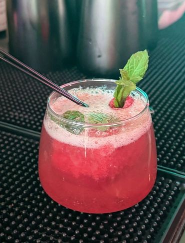 Red cocktail garnished with mint leaves and raspberry in a round glass.