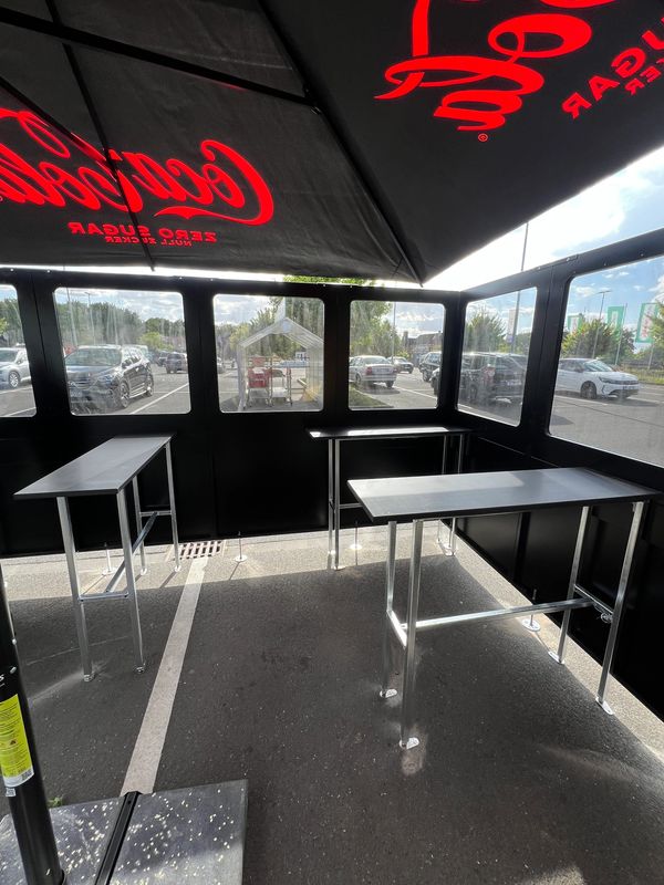 Outdoor seating area with tall tables under a Coca-Cola umbrella in a parking lot.