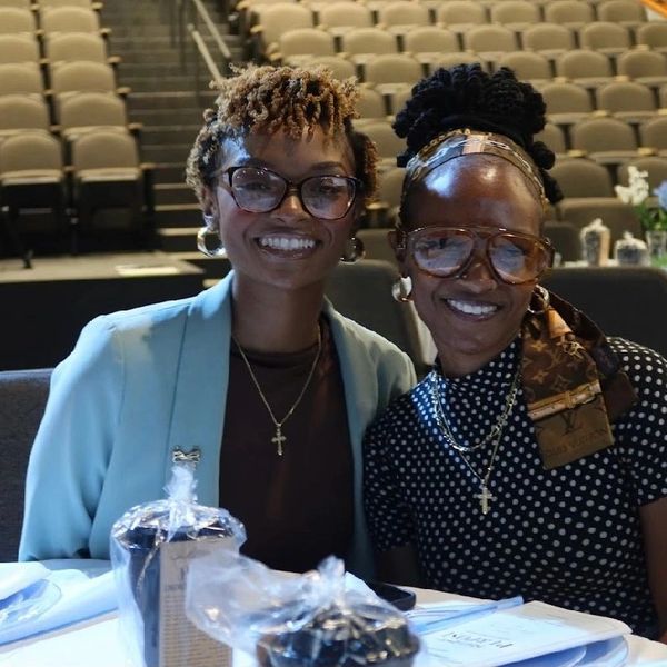 Two smiling women wearing glasses and cross necklaces seated at a table.