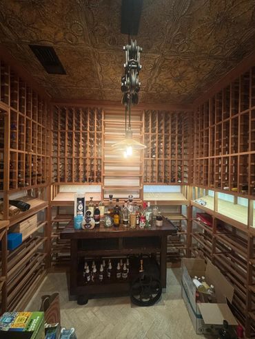 Spacious wine cellar with wooden racks and a central table displaying various liquor bottles.