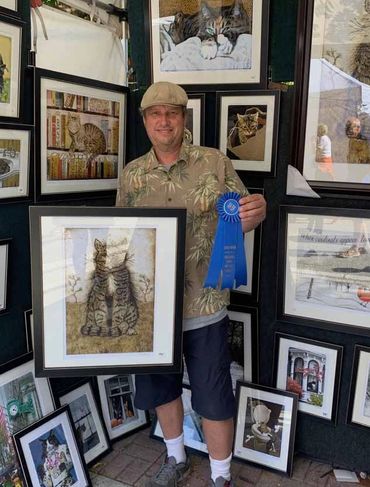 Artist proudly holding a blue ribbon and framed cat artwork at an outdoor art display.