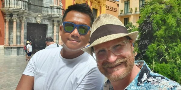 Two men smiling outdoors in a sunny urban setting.