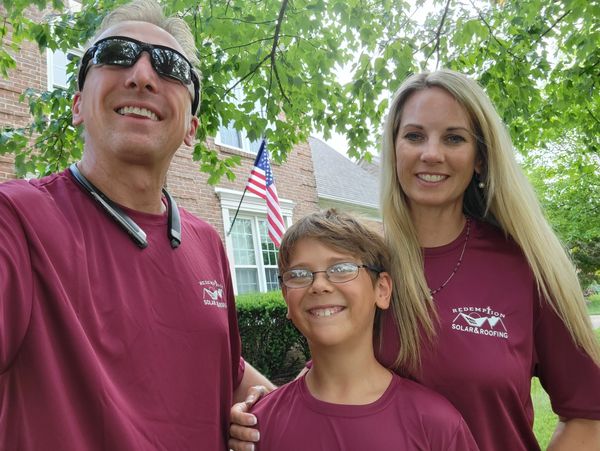 A smiling family wears matching maroon shirts outdoors under green trees.