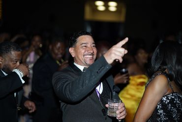 Man in black suit smiling and pointing at a lively party.