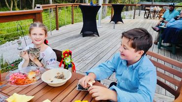 Two children sitting at an outdoor wooden table, one holding a cat and the other smiling.