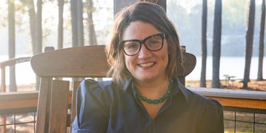 A woman in glasses sits smiling on a wooden chair outdoors.