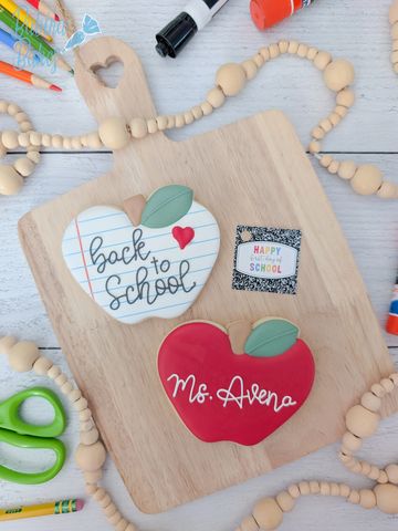 Two apple-shaped back-to-school decorated cookies on a wooden board.