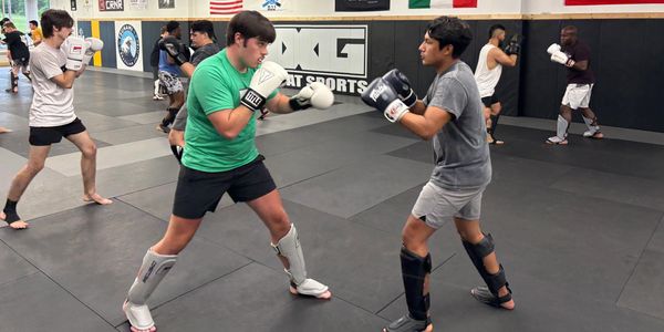 Two men sparring in a martial arts gym with others training in the background.