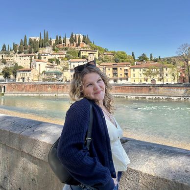 Woman posing by a riverside with hillside buildings in the background.
