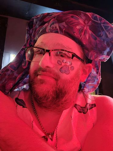 Bearded man with glasses, butterfly headscarf, and paw print face paint under red light.