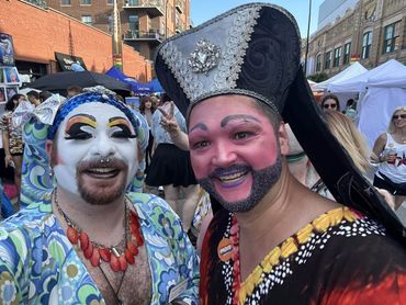 Two people in colorful, elaborate makeup and costumes at an outdoor event.