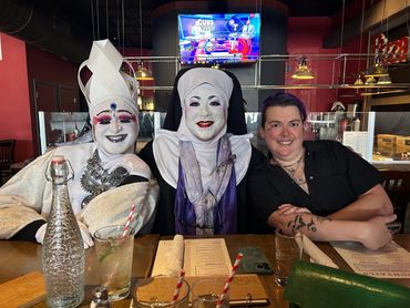 Two people in nun costumes and one person with tattoos sitting at a restaurant table.