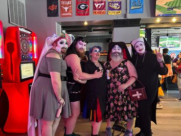Group of five people in colorful drag and nun costumes posing indoors at a bar.