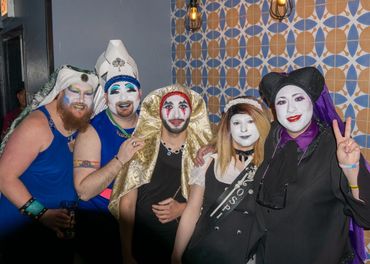 Five people in colorful, theatrical makeup and costumes posing indoors against a patterned wall.