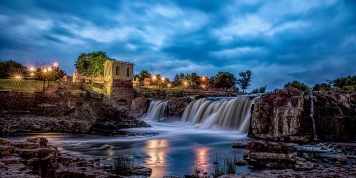 Waterfall flowing at dusk with lights and building.
