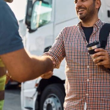 Two men shaking hands in front of a truck, one holding a coffee.