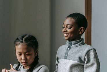 Two children collaborating, one writing in a notebook while the other watches and smiles.