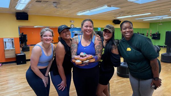Five women smiling in a gym, one holding a plate of donuts.