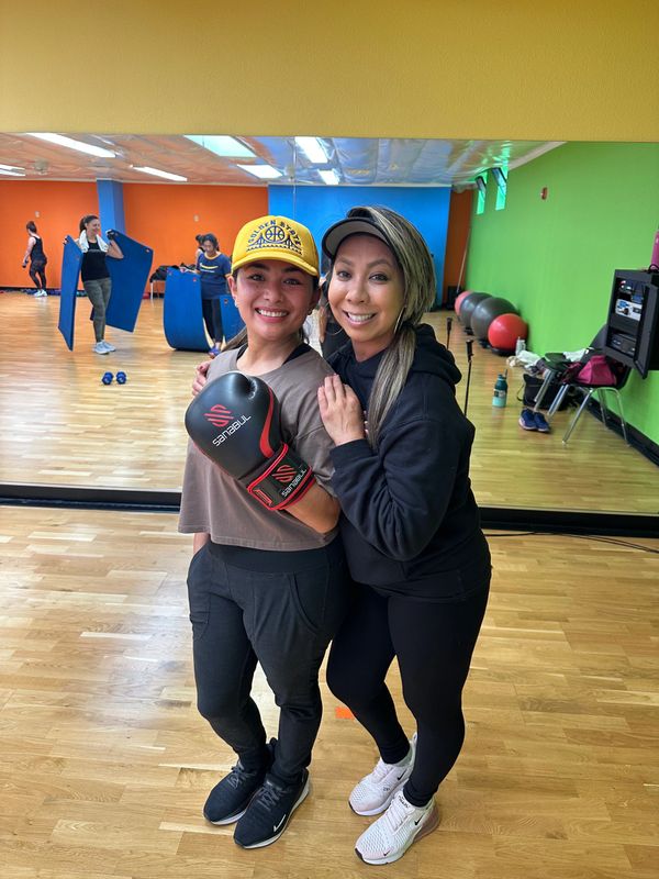 Two women smiling in a gym, one wearing boxing gloves and a yellow cap.