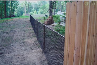 A black chain-link fence runs alongside a wooden fence in a wooded area.