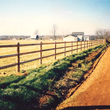 A rural dirt road alongside a wooden fence and farm buildings on a clear day.