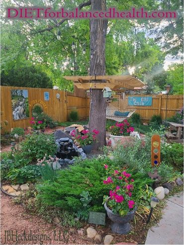 Backyard landscape with pink flowers and green shrubs, water fountain, lots of art and hammock.